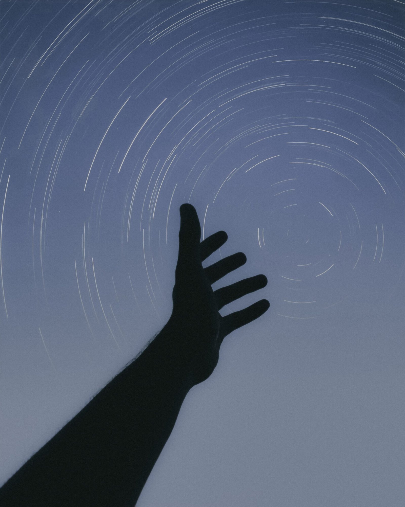 Home Silhouette of a hand reaching upward against a stunning star trail background in a night sky.